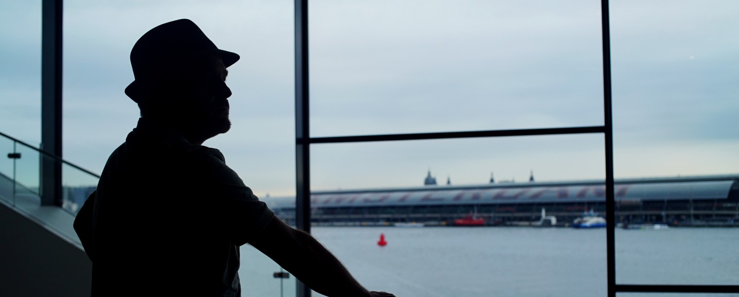 Silhouette van Dirk met hoed die uit raam kijkt over 't IJ naar Amsterdam Centraal